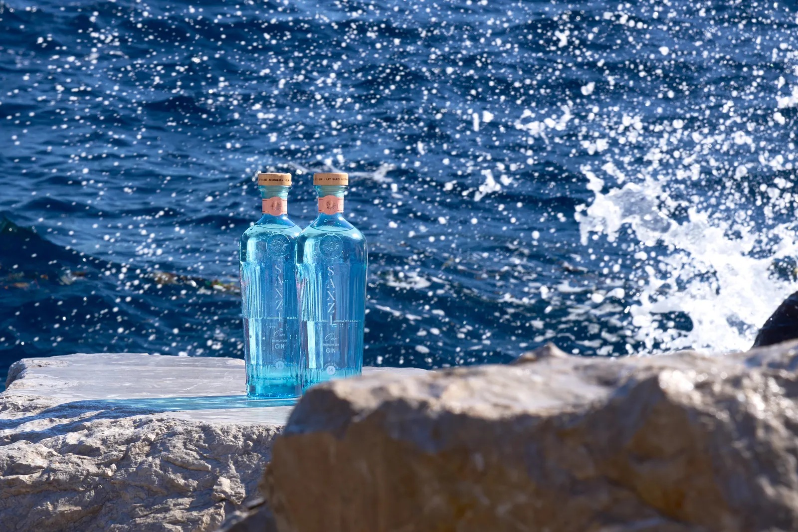 Two bottles of Saxzi One gin on a stone ledge above the Mediterranean